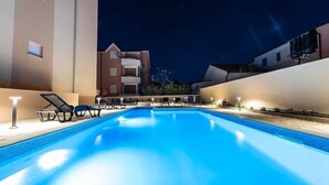 A swimming pool with lounge chairs and lights, surrounded by buildings at night.