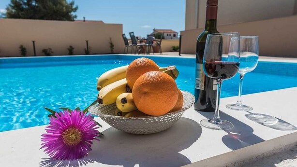 Une table placée au bord de la piscine, avec un saladier rempli de fruits, une bouteille de vin et deux verres.
