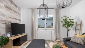 Una sala de estar moderna con un televisor de pantalla plana sobre un mueble de madera, un ventanal con cortinas, un sofá negro, una mesa de centro con objetos decorativos y una planta en maceta.