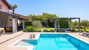 Una piscina con pérgola, mobiliario exterior y una zona de patio con mesa y sillas.