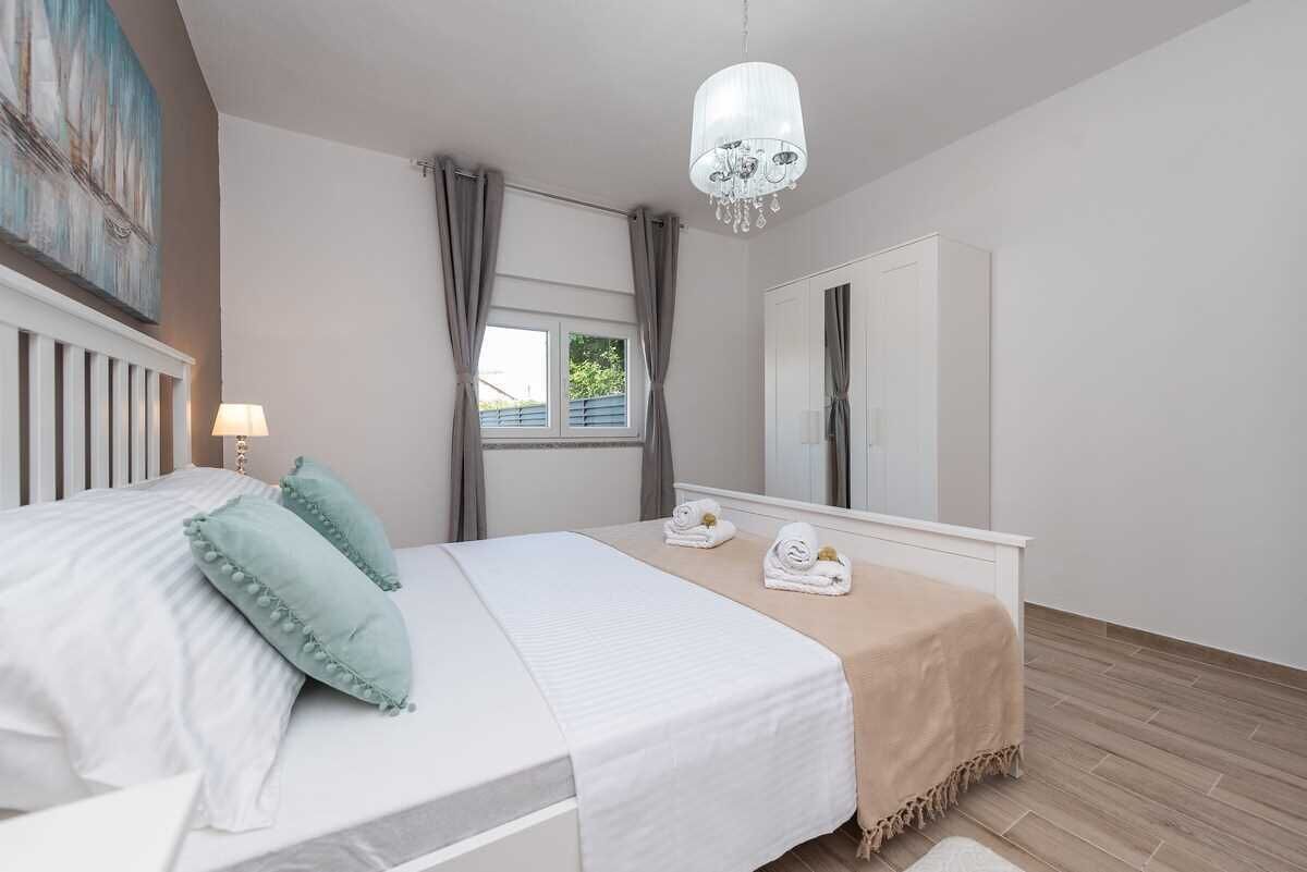 A bedroom with a bed, a chandelier, a window with curtains, a wardrobe, and a painting on the wall.