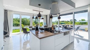 Une cuisine moderne dotée d’un grand îlot central, de luminaires suspendus et offrant une vue sur la verdure environnante.