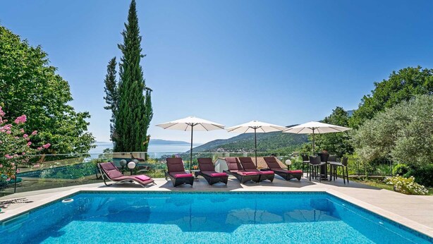 Piscina con sillas de descanso y sombrillas, rodeada de vegetación y vistas al mar.