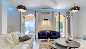 Una sala de estar moderna con un sofá de cuero blanco, dos sillones oscuros y una mesa de centro redonda. Amplios ventanales ofrecen vistas al exterior.