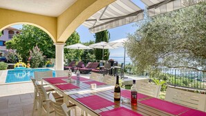 Zona junto a la piscina con mesa y sillas, sombrillas y vistas al mar.