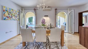 Comedor con mesa dispuesta para cuatro personas, una araña de cristal y vistas al exterior a través de amplios ventanales.