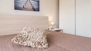 A bedroom with a wooden headboard, a pink bedspread, a textured throw pillow, a nightstand with a lamp, and a painting of a pier on the wall.