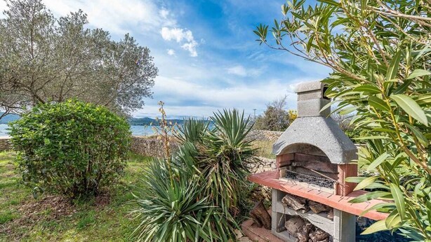 A stone outdoor grill with a chimney, surrounded by lush greenery and overlooking a body of water.