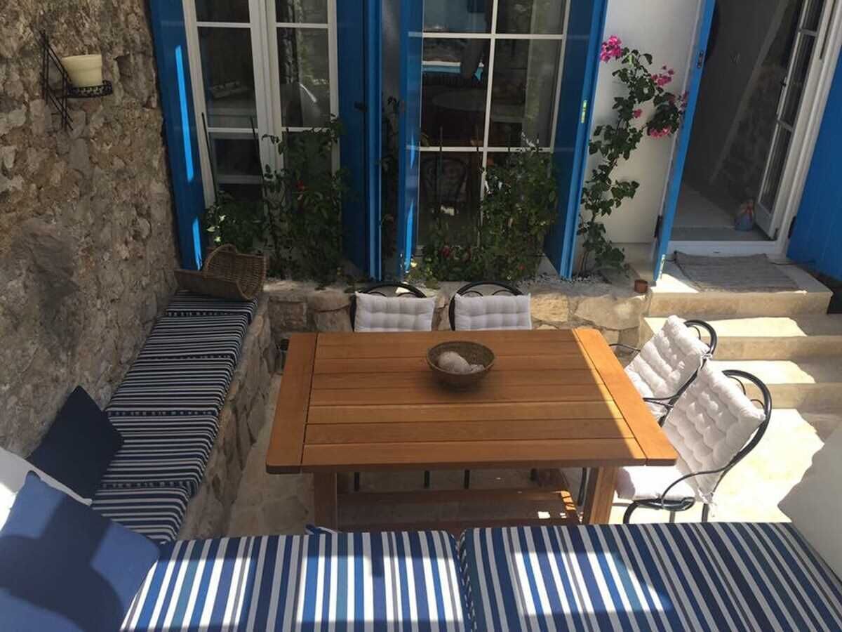 A wooden table with a bowl, surrounded by striped cushions and white chairs, set against a stone wall with a blue door and potted plants.
