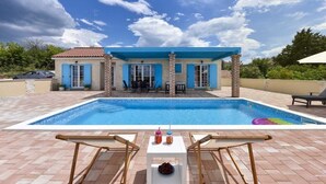Una piscina con borde de azulejos azules, dos sillas de descanso y una mesa con bebidas y aperitivos.