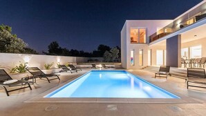 A modern outdoor pool area with lounge chairs and a well-lit building with a balcony. - Villa Pollux | Adriatic Luxury Villas (Vrsi)