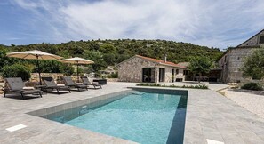 A pool area with lounge chairs and umbrellas, a stone building, and a hillside in the background.