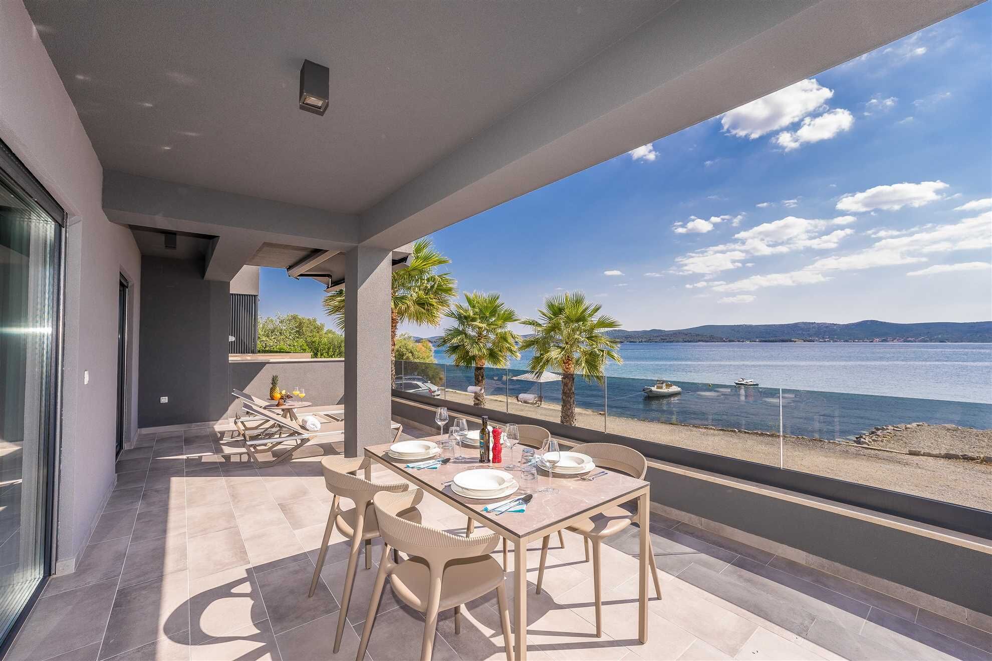 A balcony with a table set for dining, overlooking a calm sea with boats and palm trees.