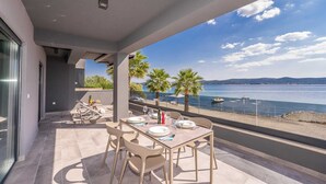 A balcony with a table set for dining, overlooking a calm sea with boats and palm trees.