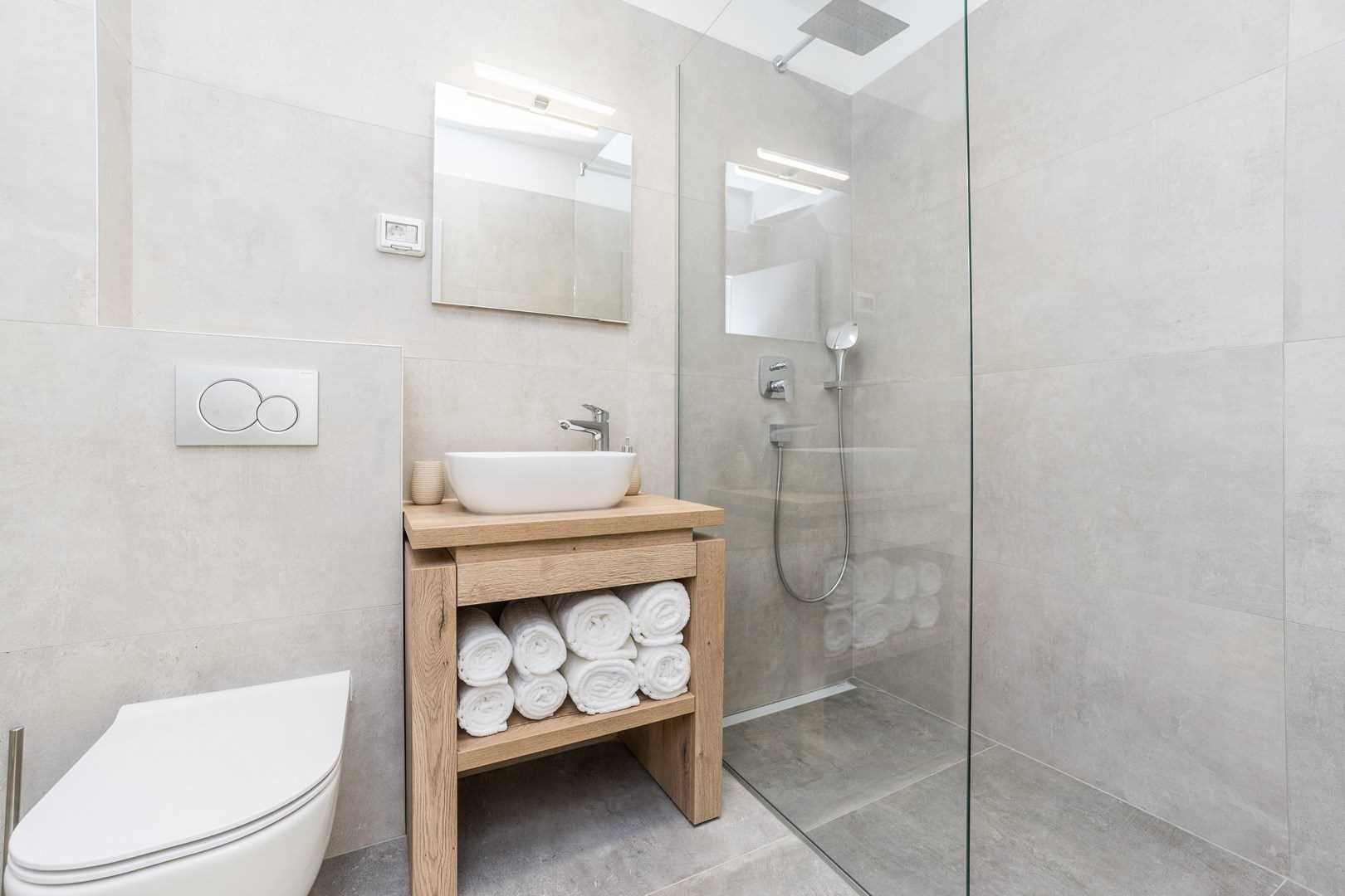 Et moderne badeværelse med vask i hvidt, trækommode, glas brusekabine og toilet.