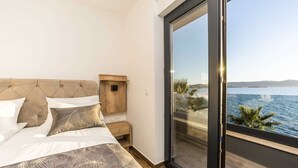 A bedroom with a bed, a nightstand, a wooden cabinet, and a large sliding glass door leading to a balcony with a view of the sea.