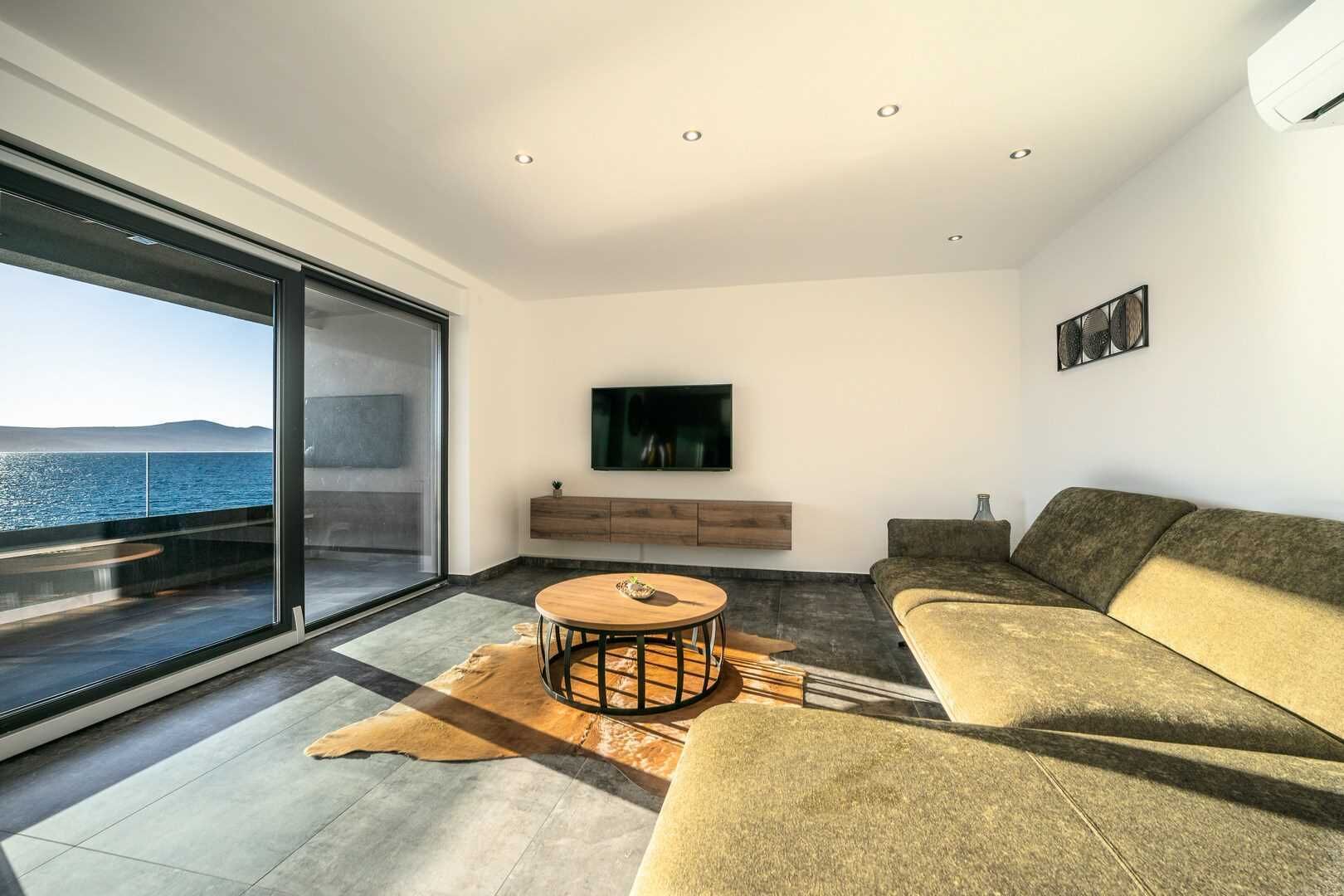 Una sala de estar moderna con un gran ventanal corredero, una mesa de centro redonda de madera, un sofá, un televisor montado y un mueble de madera minimalista.