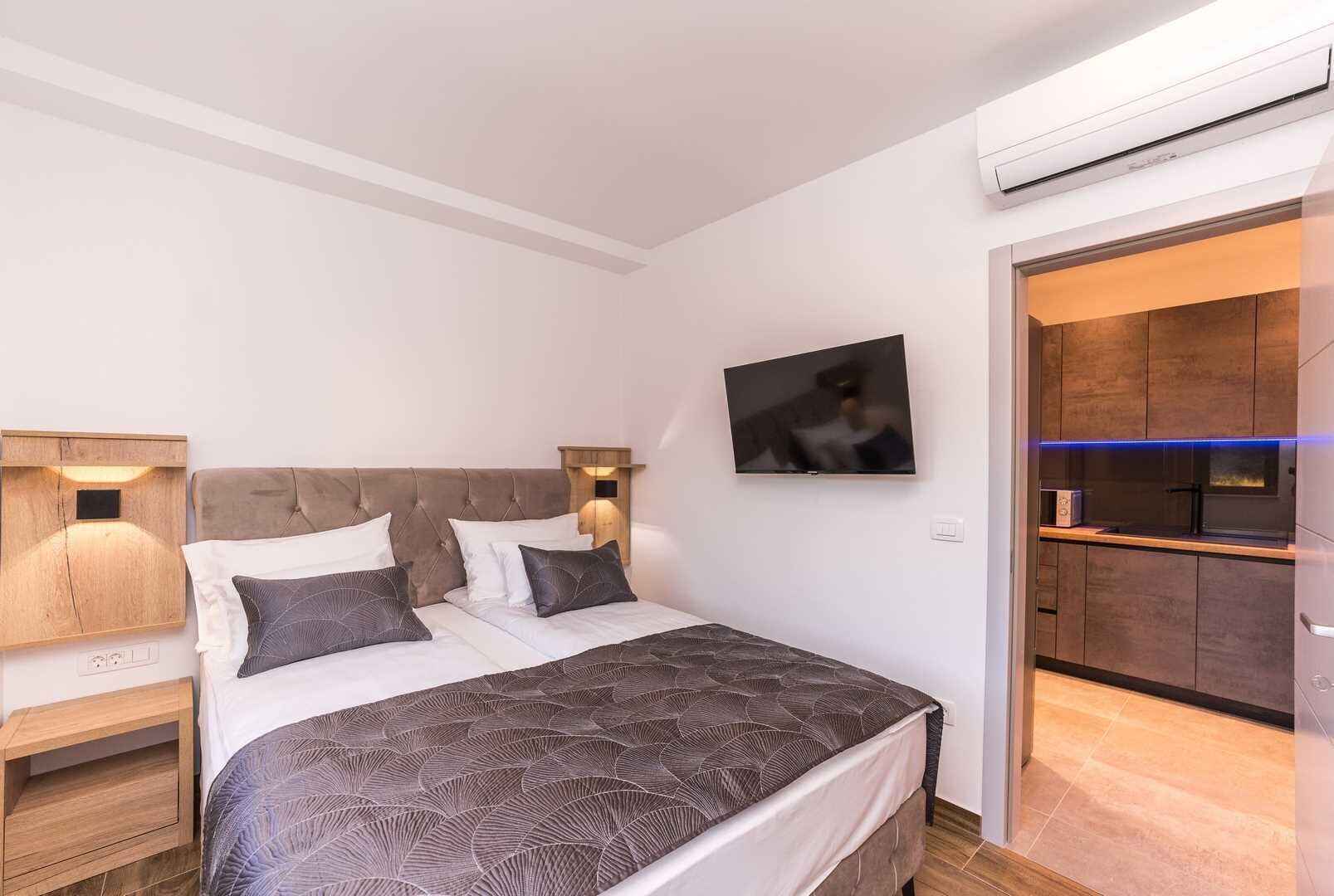 A bedroom with a bed, bedside tables, a TV, and a view into a kitchen area.