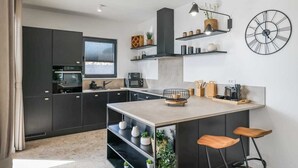 A modern kitchen with dark cabinetry, a central island with bar stools, built-in shelves with decorative items, and a large wall clock. - Villa Dekorti | Adriatic Luxury Villas (Kožino)
