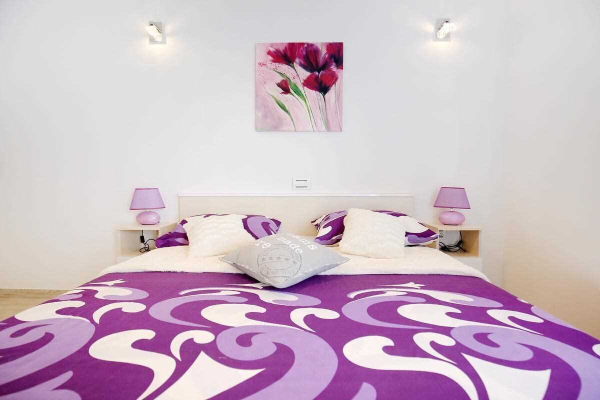 A neatly made bed with a purple and white patterned bedspread, two bedside lamps, and a wall-mounted painting of red flowers.