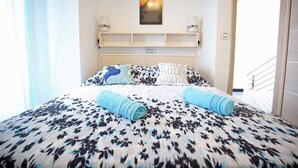 Habitación de hotel con una cama cubierta con un edredón floral en blanco y negro, dos toallas enrolladas, un estante de pared con dos lámparas y una cita inspiradora enmarcada sobre la cama.