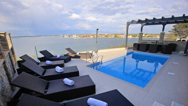 Un área junto a la piscina con sillones de descanso y una pérgola con vista a un cuerpo de agua.