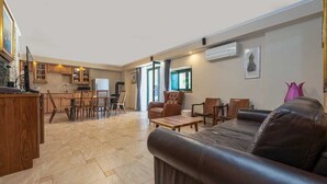 A spacious living area with a leather sofa, wooden dining table, and chairs. There is a kitchen area with wooden cabinets and a refrigerator.