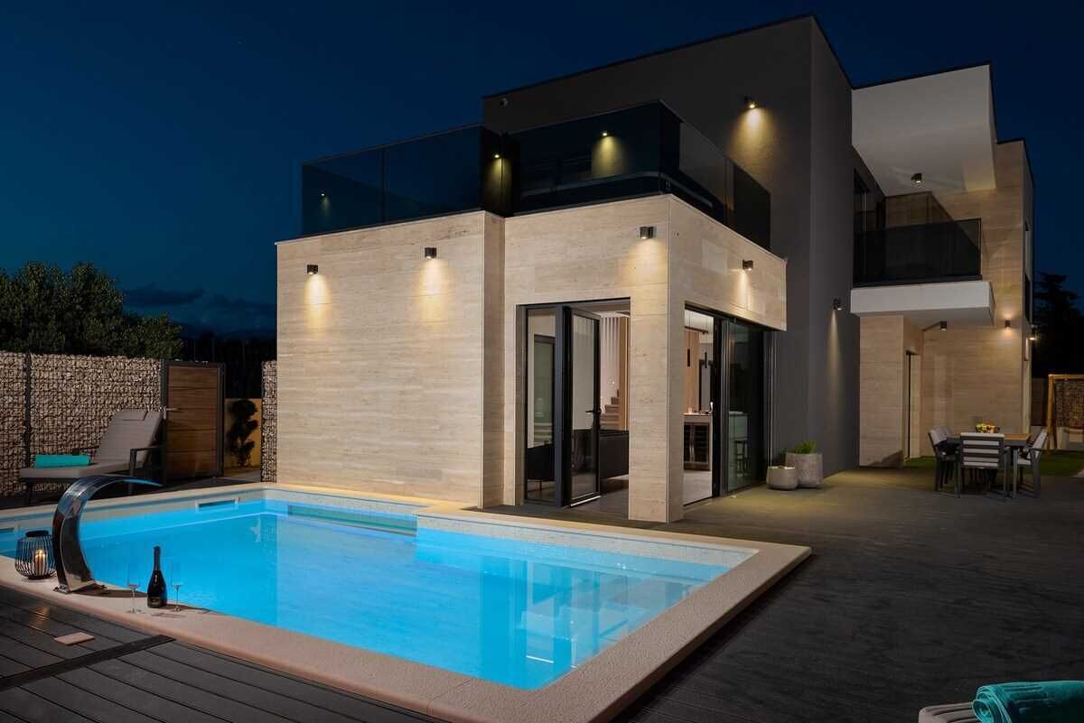A modern two-story house with a swimming pool, outdoor seating, and a well-lit patio area.