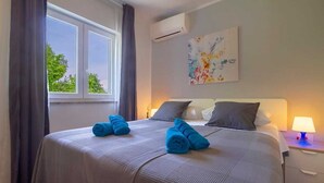 A hotel room with a large bed, a nightstand with a lamp, a window with curtains, and a wall-mounted air conditioner. - Villa Meline | Adriatic Luxury Villas (Donja Hlapa)