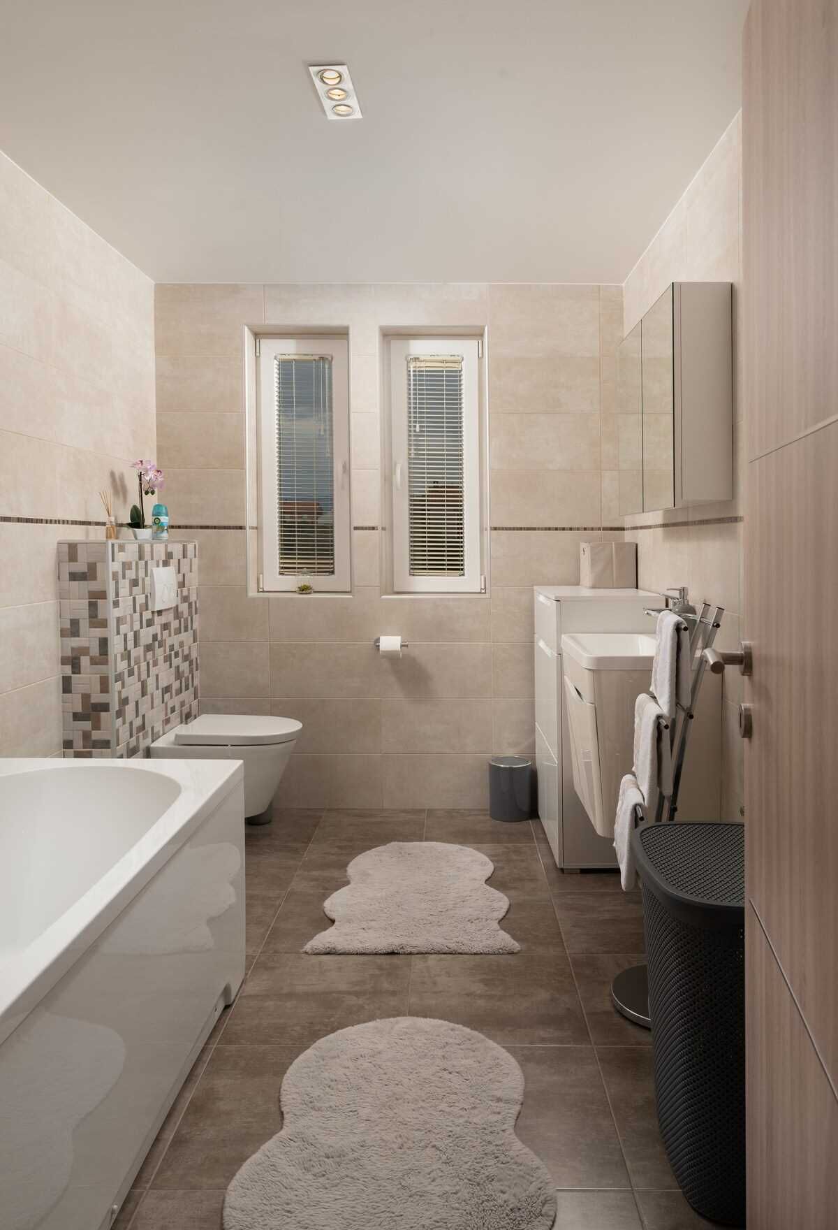 Un bagno moderno con vasca, water e lavatrice. Due finestre con persiane, un rivestimento in piastrelle e un piccolo tappeto sul pavimento.