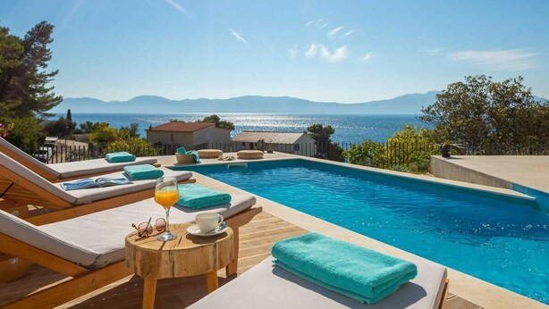 Zona junto a la piscina con sillones, una mesa con una bebida y vistas al mar y a las montañas.
