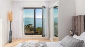 Un dormitorio con cama, una mesita de noche, un florero y vista al mar a través de una puerta corrediza de vidrio.