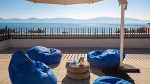 Una terraza con vistas al mar y a las montañas, con puffs azules y una cesta tejida que contiene un libro y una taza.