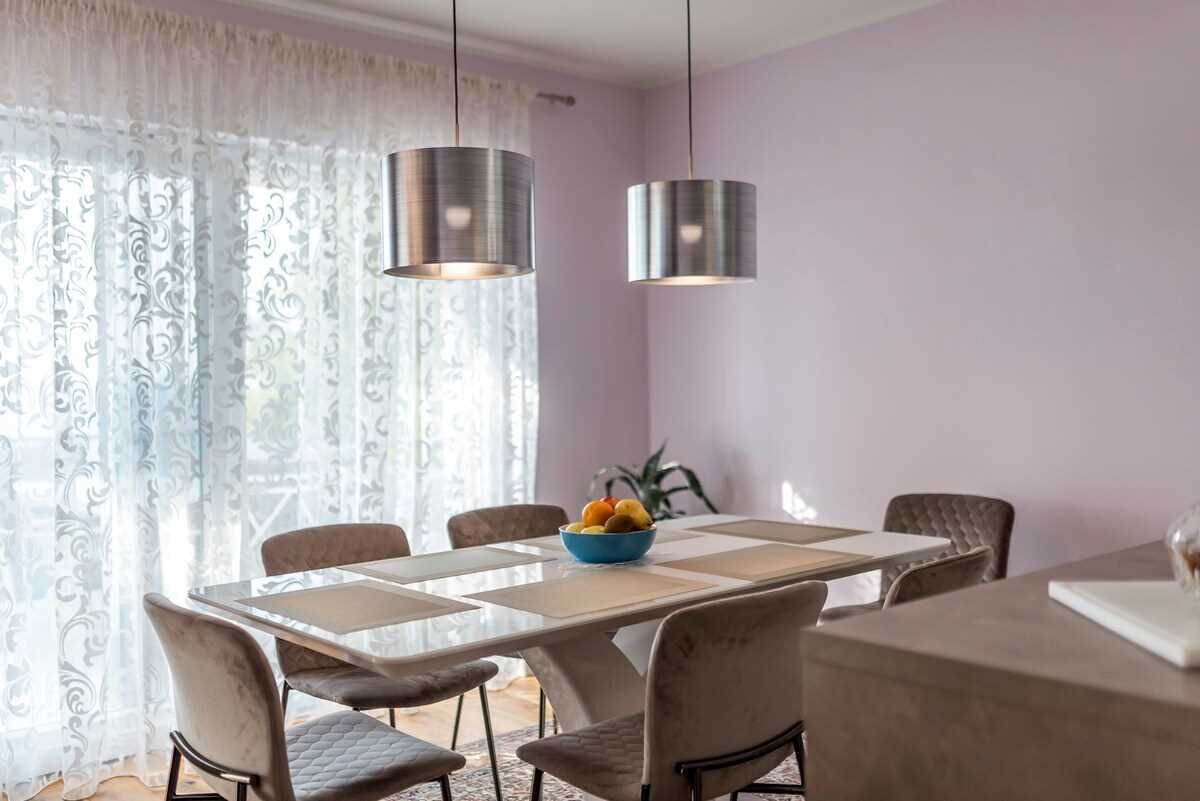 Un coin repas avec une table dressée pour six personnes, deux lampes suspendues et une fenêtre ornée de rideaux légers.