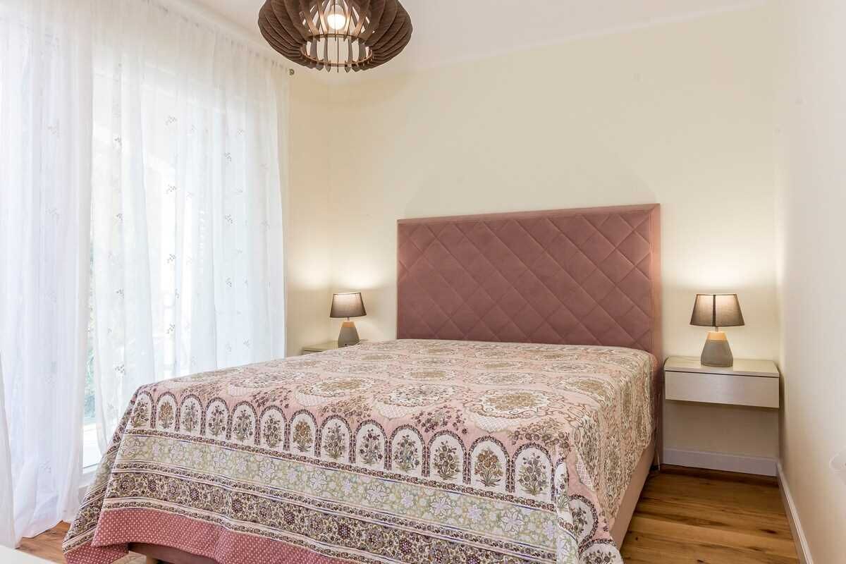 A bedroom with a quilted headboard, two bedside tables with lamps, and a window with sheer curtains.