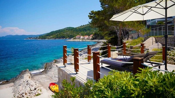 A coastal hotel with a lounge area overlooking the sea, a rocky shoreline, and a view of the distant landscape.