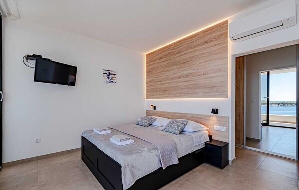 Un dormitorio moderno con cama, mesitas de noche, televisor montado y vista a la playa a través de un ventanal.