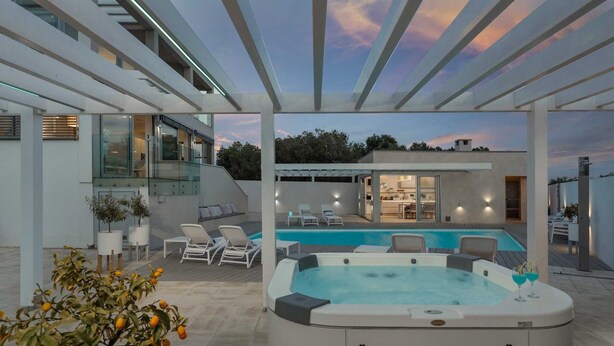 A modern outdoor pool area with a hot tub, lounge chairs, and a glass-enclosed kitchen.