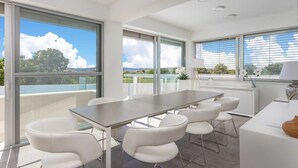 A modern dining area with a long table and six white chairs, large windows with a view of the outdoors, and a white countertop with a lamp.