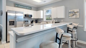 Una cocina moderna con armarios blancos, una isla grande con taburetes de bar, un refrigerador de acero inoxidable y un letrero que dice 'EAT. BEACH. SLEEP. REPEAT'.