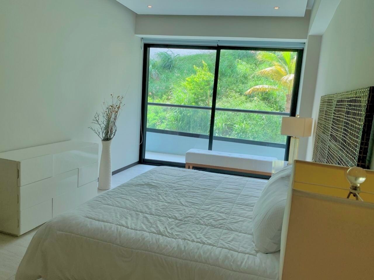 A modern bedroom with a large window, a bed, a bedside table, and a white dresser.