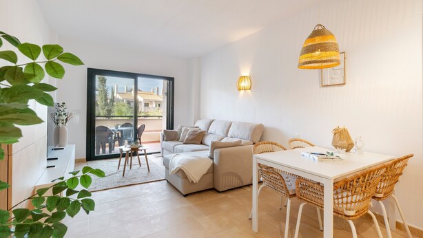A modern living room with a sofa, dining table, wicker chairs, a pendant light, and a view through a glass door.