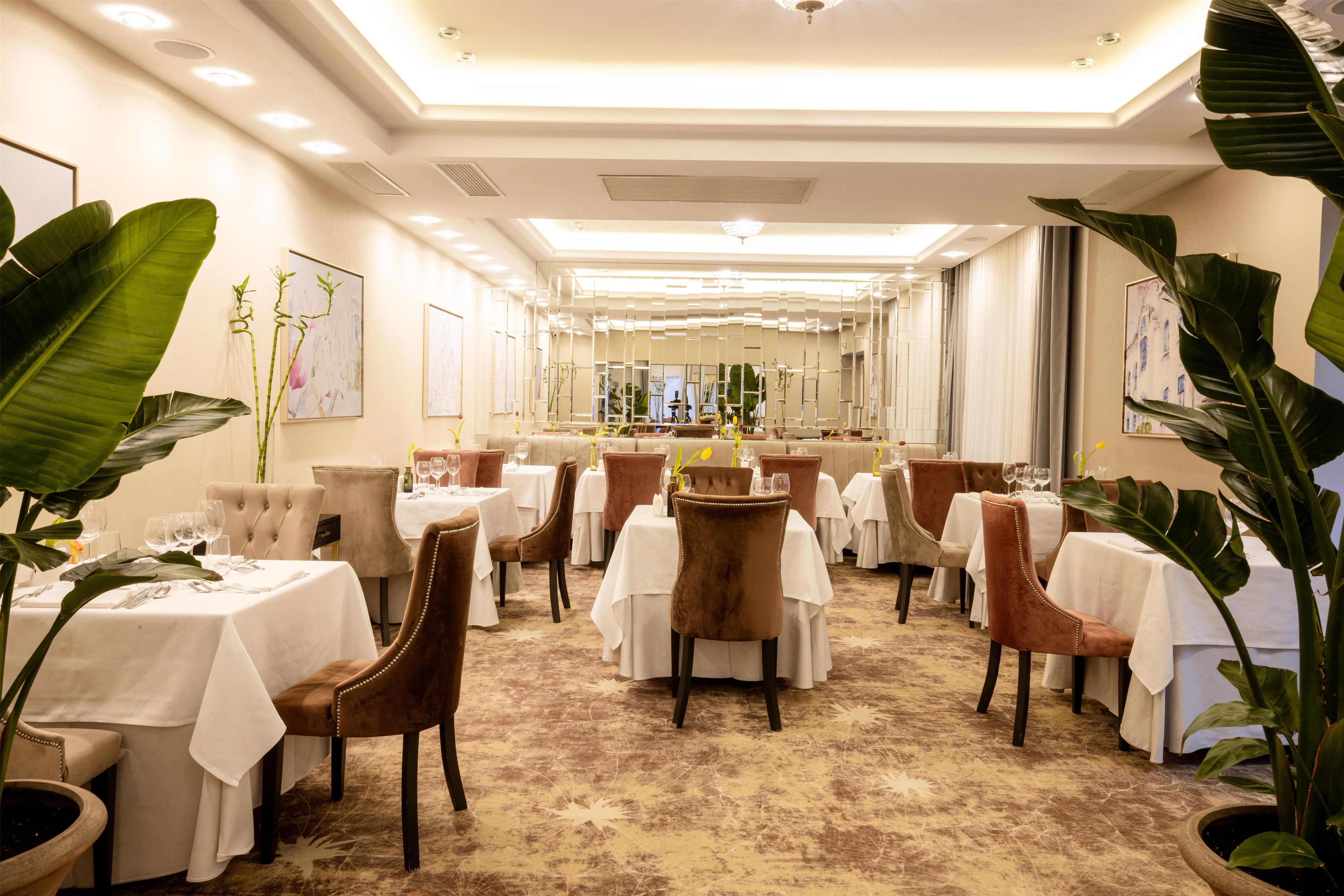 A hotel dining area with multiple tables and chairs, a patterned carpet, and large potted plants.