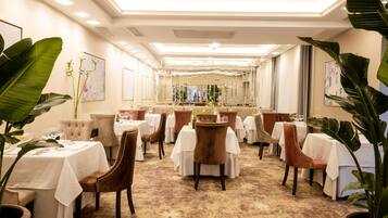 A hotel dining area with multiple tables and chairs, a patterned carpet, and large potted plants.
