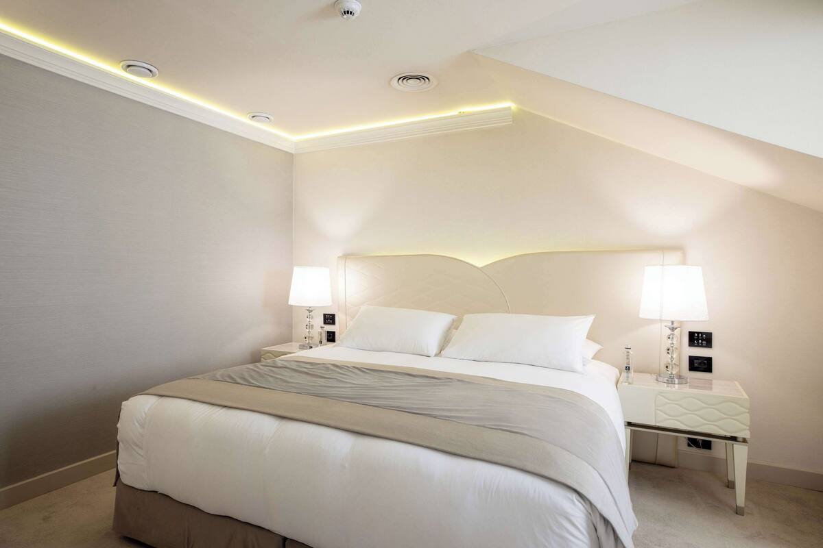 Une chambre d’hôtel moderne avec un grand lit, deux tables de chevet avec des lampes et un plafond doté d’un éclairage encastré.