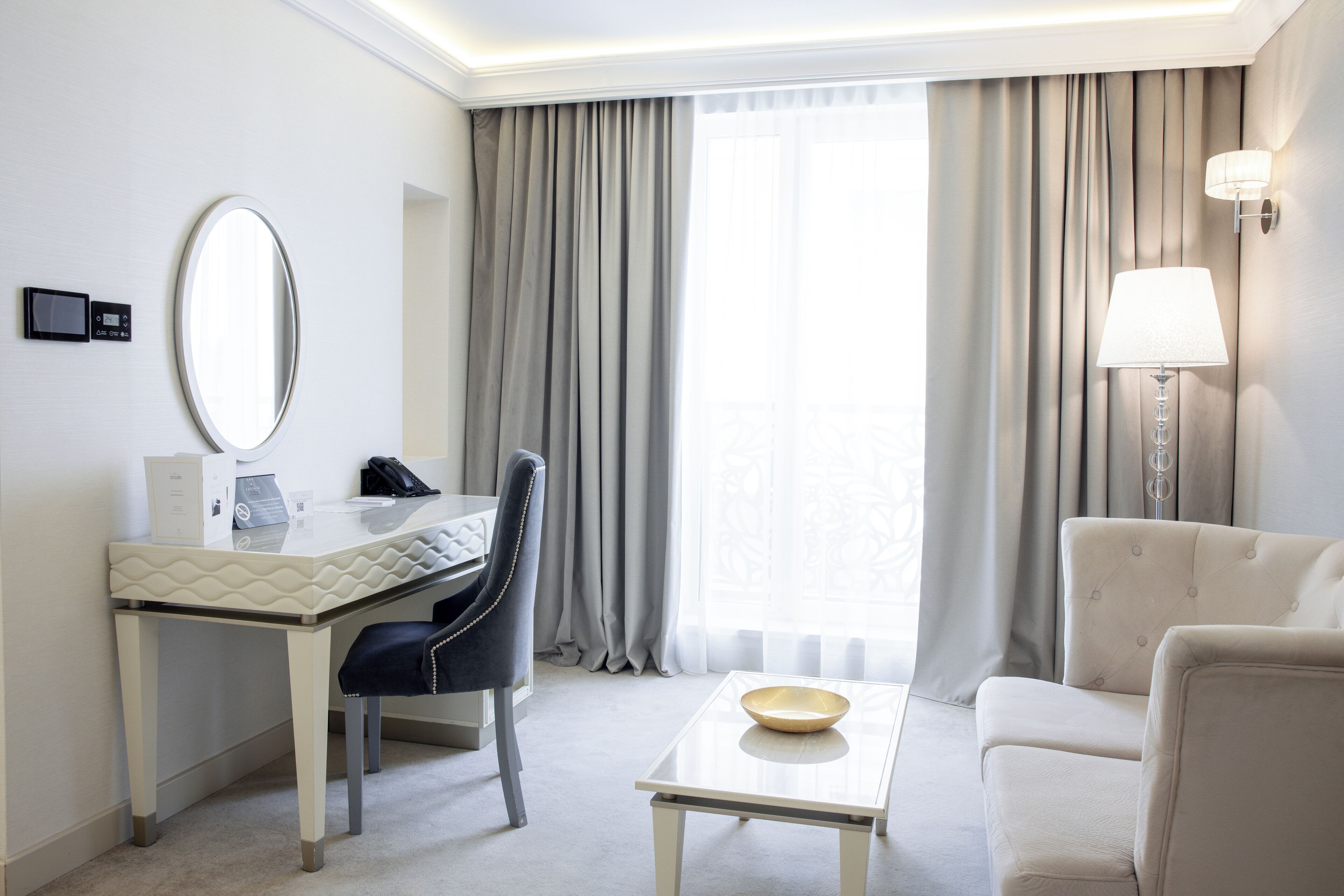 Ein Hotelzimmer mit Schreibtisch, Stuhl und einer Couch. An der Wand hängt eine Lampe und es gibt ein Fenster mit Vorhängen.