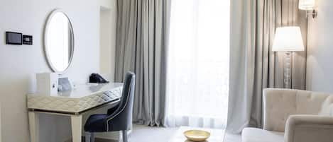 A hotel room with a desk, chair, and a sofa. There is a lamp on the wall and a window with curtains.