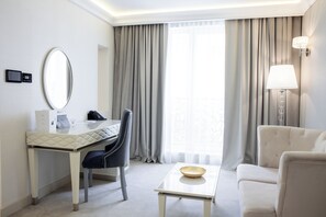 Habitación de hotel con escritorio, silla y sofá. Hay una lámpara en la pared y una ventana con cortinas.
