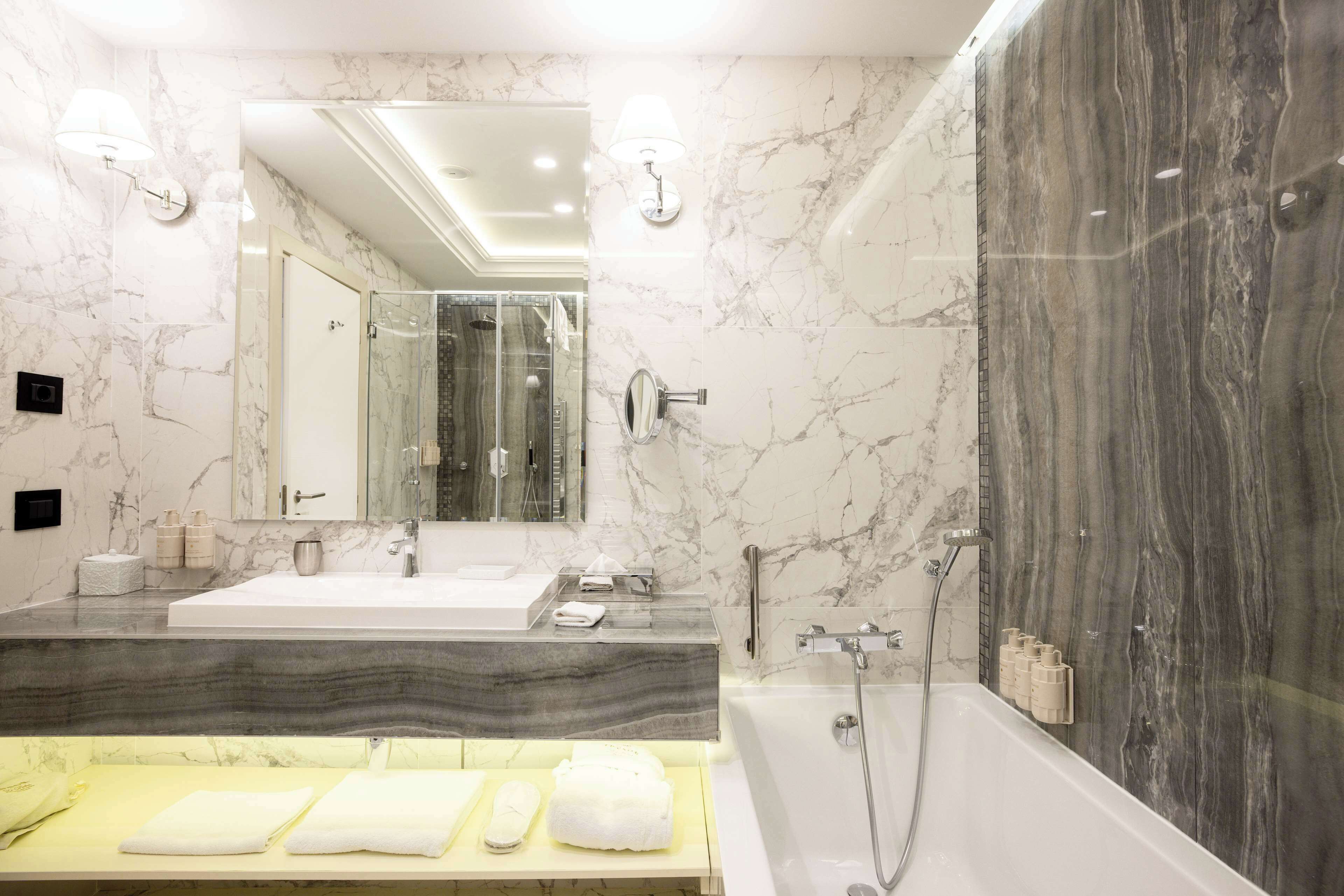 Une salle de bain moderne avec des murs en marbre, un grand miroir, une baignoire et un meuble-lavabo agrémenté de plusieurs serviettes.