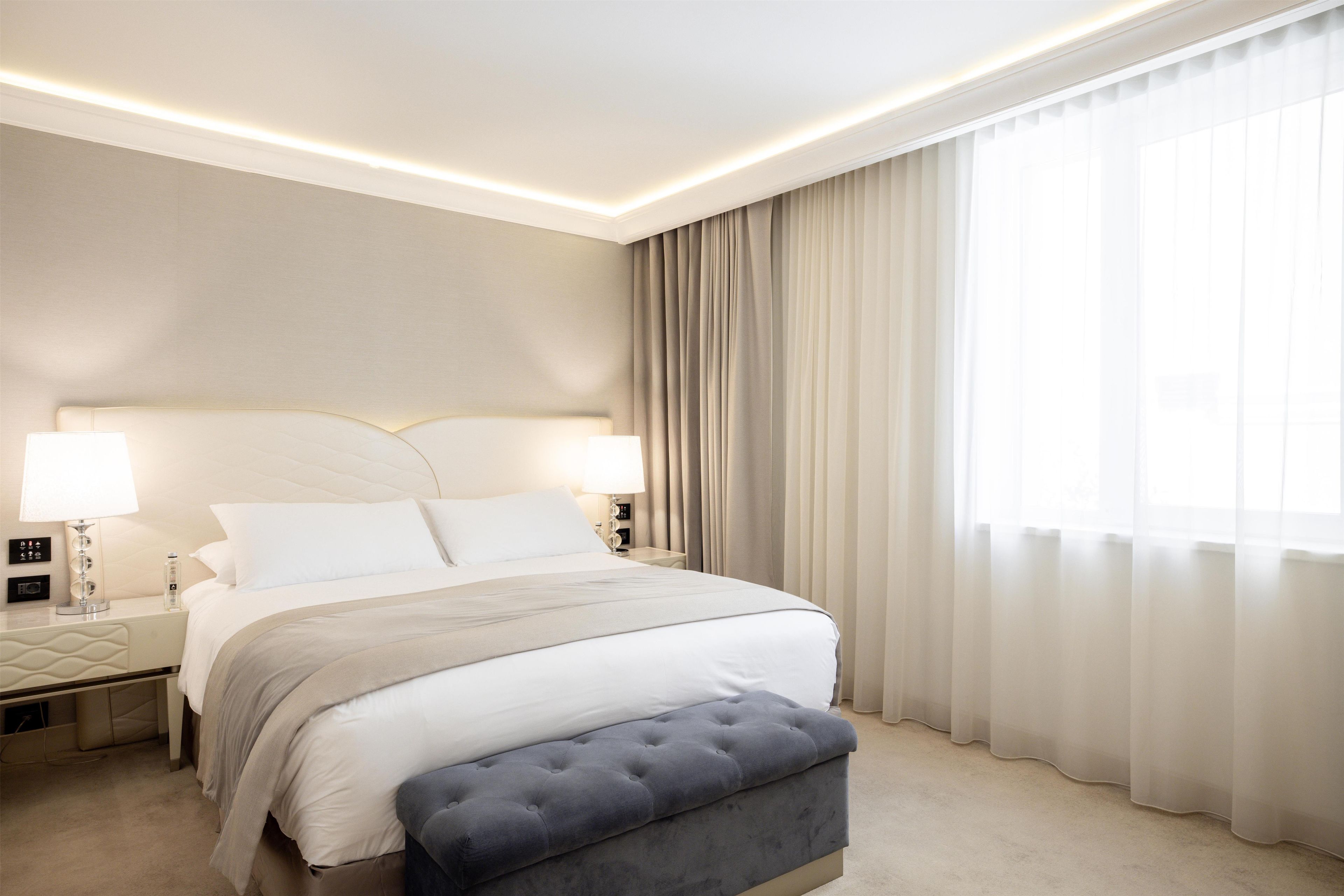 Una habitación de hotel con cama, lámparas de noche, mesita de noche, ventana con cortinas y piso alfombrado.